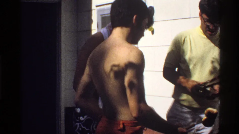 1978 California frame showing men in swimwear and casual clothing gathered in a tiled private room at a backyard party.