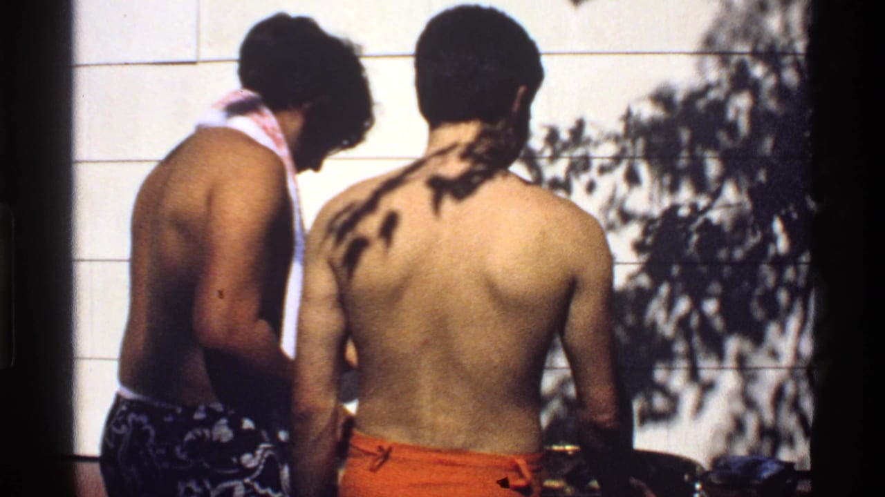 Two shirtless young men stand by a grill in front of white siding with tree shadows.