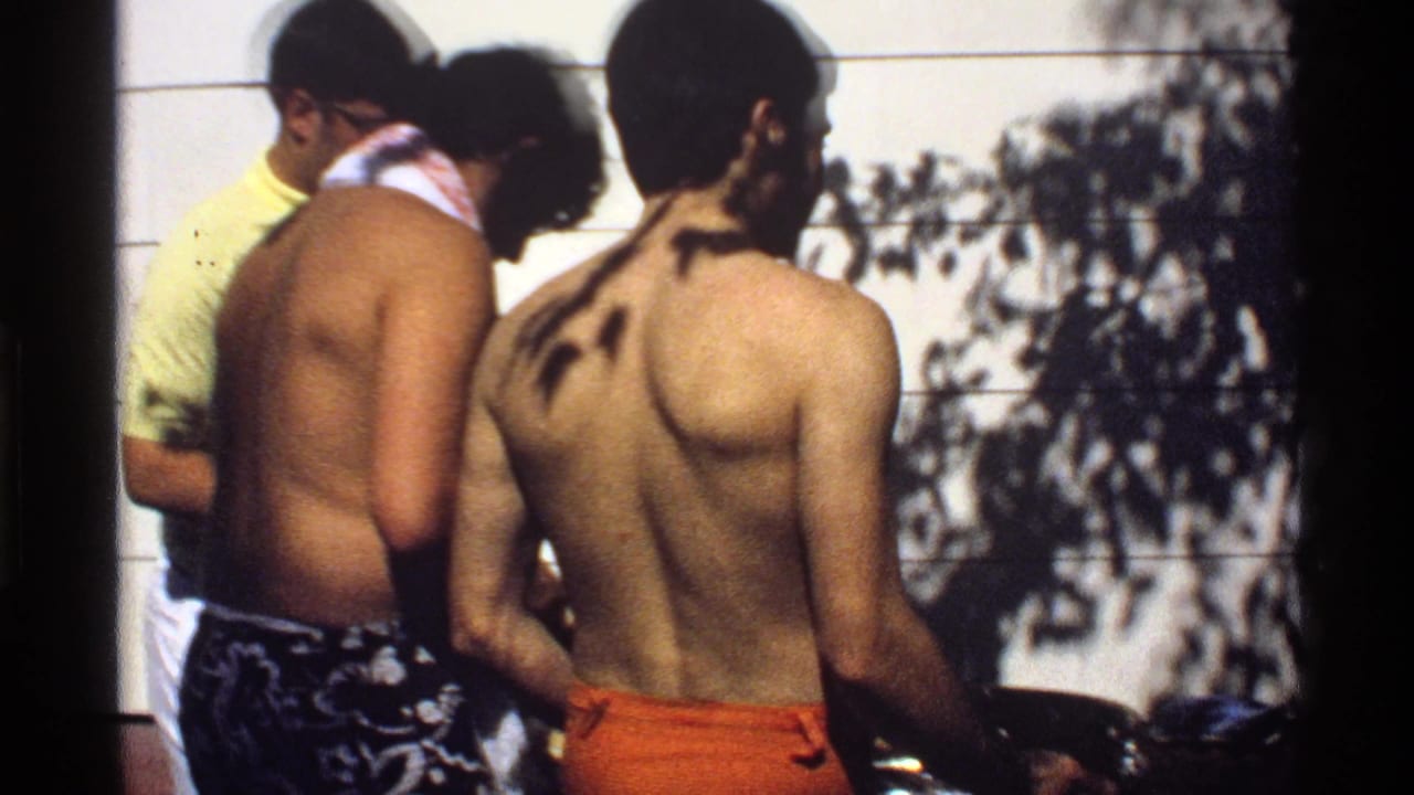 Three men gather close to a backyard grill, with tree shadows on the wall behind them.