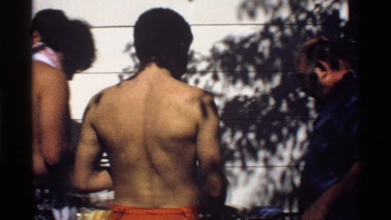 1978 California backyard cookout frame with shirtless men near the grill and an older man in a blue shirt entering the shot.