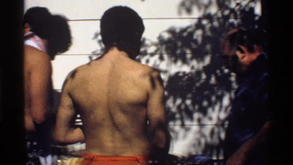 1978 California backyard cookout frame with shirtless men near the grill and an older man in a blue shirt entering the shot.
