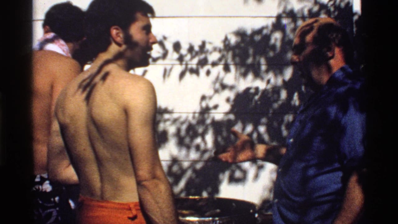 California 1978 home-movie frame showing a shirtless young man turning as an older man in a blue shirt gestures near the backyard grill.