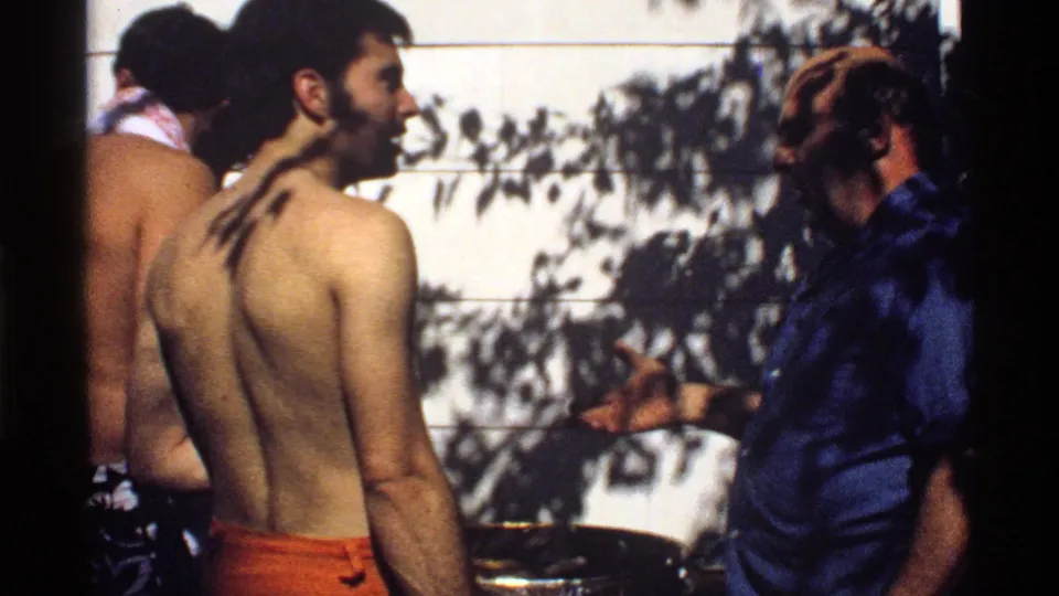 California 1978 home-movie frame showing a shirtless young man turning as an older man in a blue shirt gestures near the backyard grill.