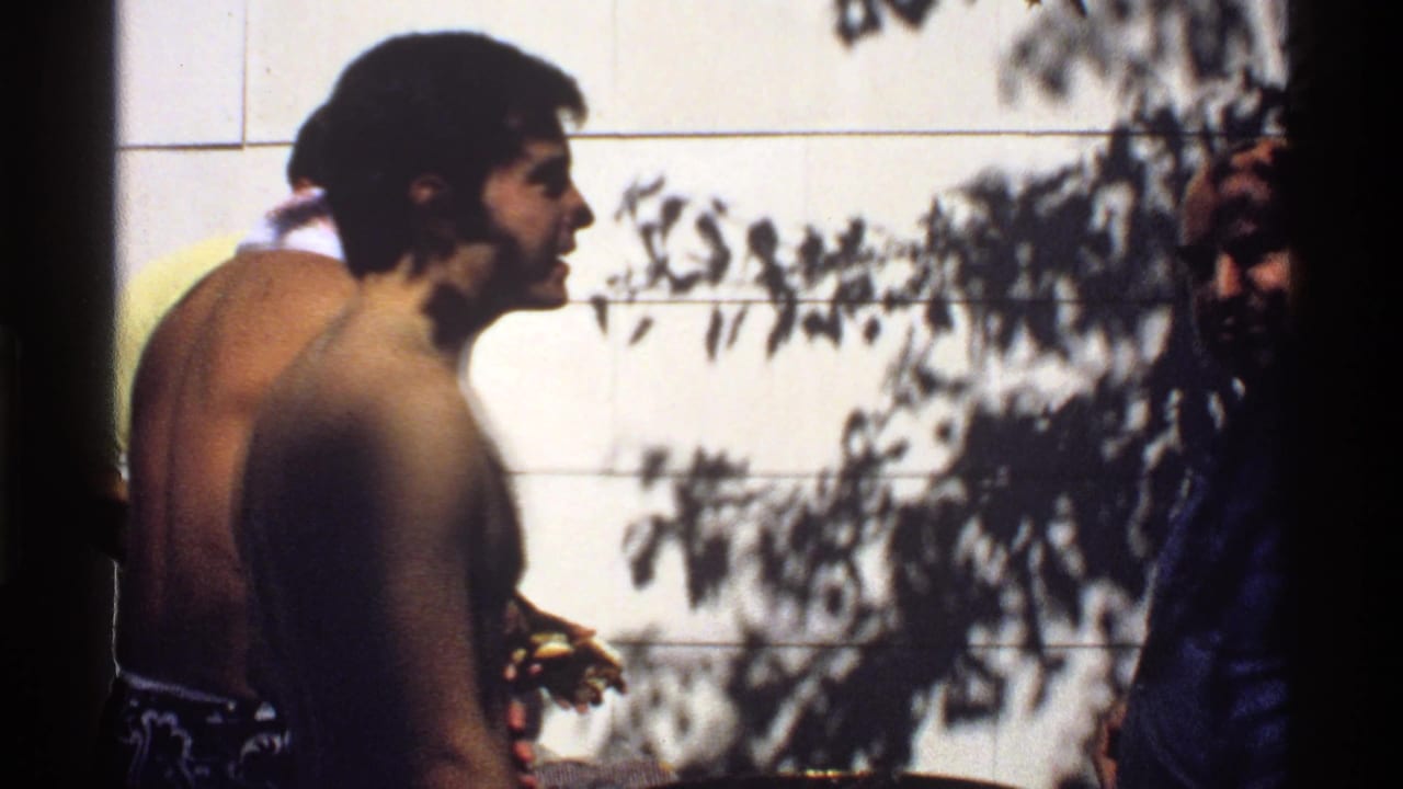 Two shirtless young men in swim trunks stand near a sunlit tiled wall with tree shadows.