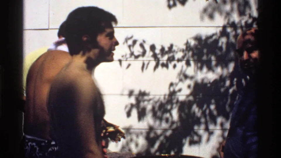 1978 California 8mm home-movie frame showing young men at poolside beside a white tiled wall with dappled tree shadows.
