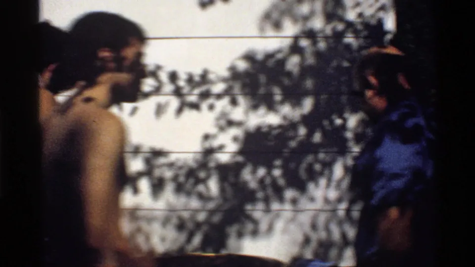 Poolside home-movie frame in California showing several young men near a tiled wall, partly obscured by shadows and frame edges.