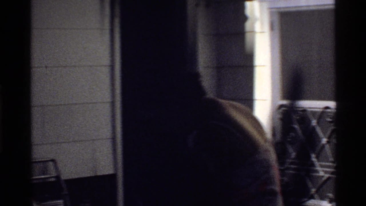 Dim 8mm frame from the poolside area showing a doorway, wall tiles, patio furniture, and a partially visible person.