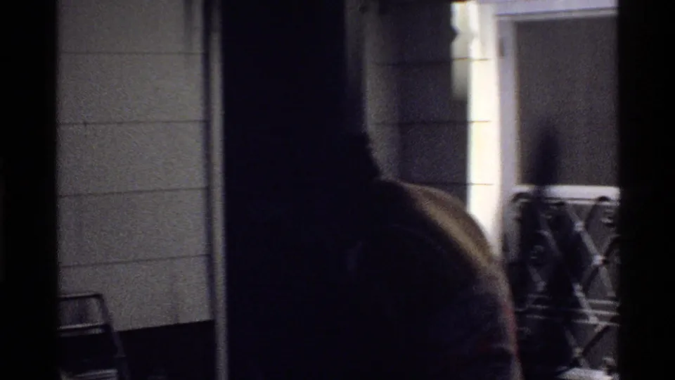 Dim 8mm frame from the poolside area showing a doorway, wall tiles, patio furniture, and a partially visible person.