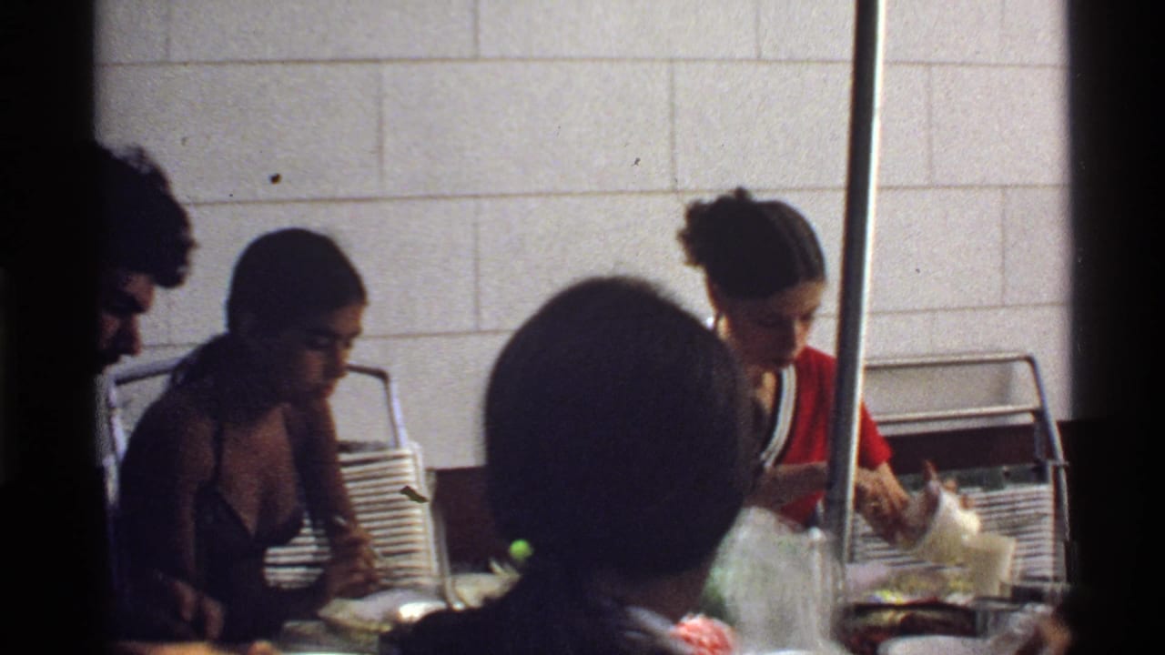 1978 California frame showing a woman in a red-and-white shirt serving food beside seated adults at an indoor table.