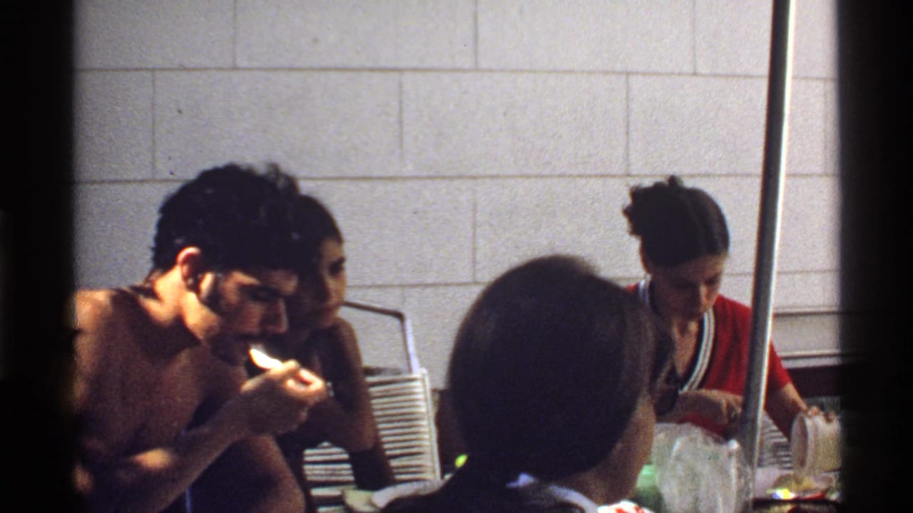 1978 California home-movie frame with a shirtless man lighting a cigarette near seated guests and tableware.