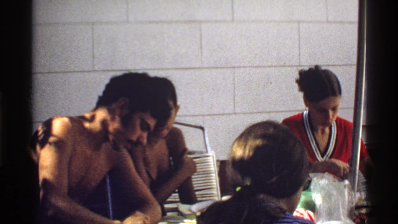 1978 California frame of adults eating and serving food at an indoor table, partly blocked by foreground heads and a dark divider.