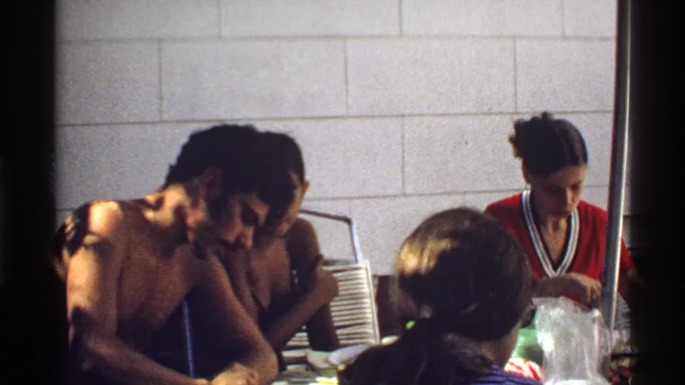1978 California frame of adults eating and serving food at an indoor table, partly blocked by foreground heads and a dark divider.
