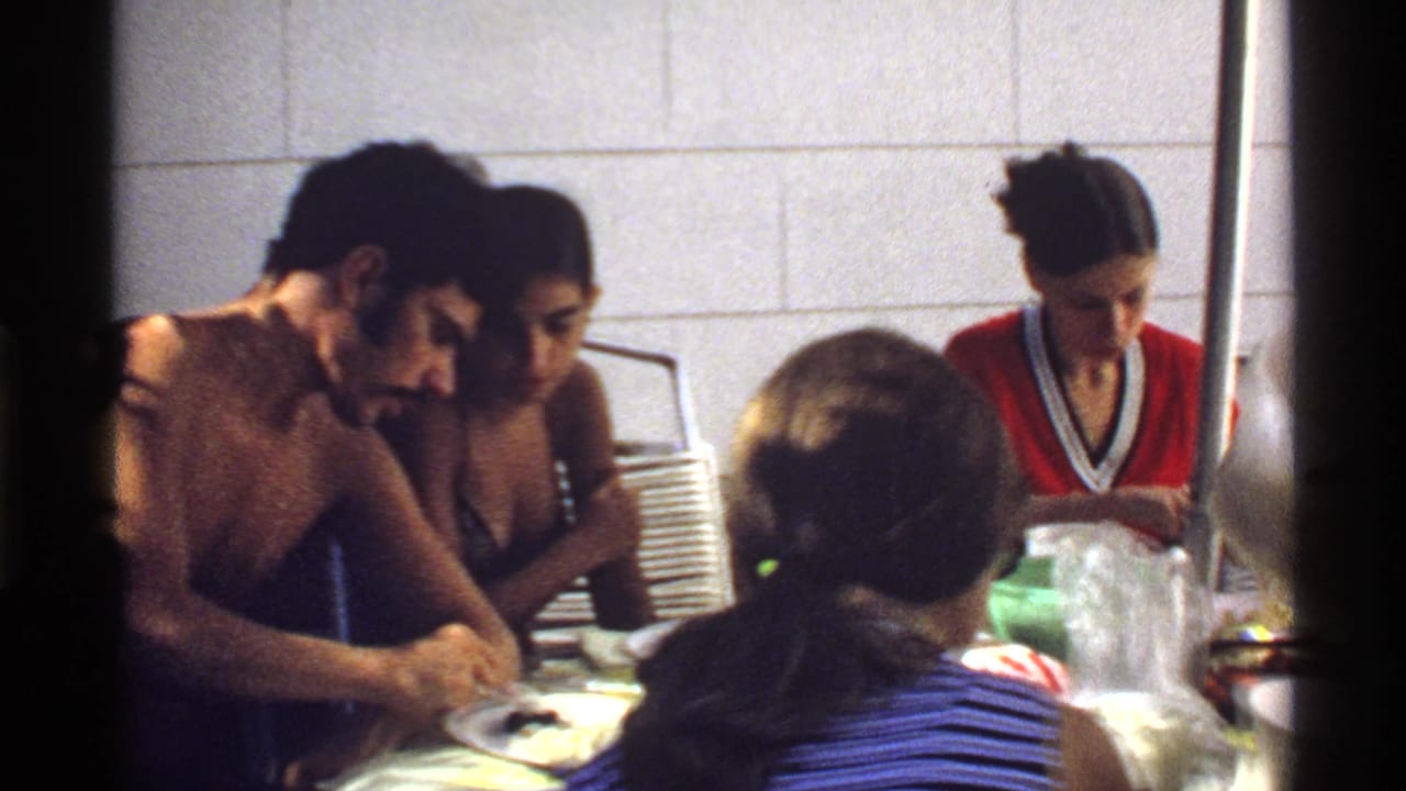 1978 California home-movie frame of a shirtless man and women gathered around a meal at an indoor table.