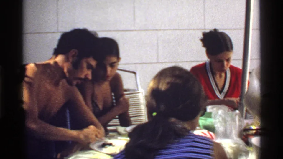 1978 California home-movie frame of a shirtless man and women gathered around a meal at an indoor table.