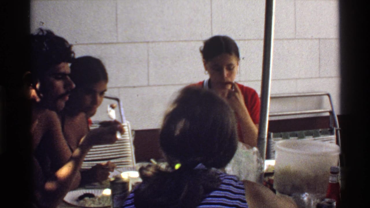 Home-movie frame from 1978 California showing young adults conversing and eating around a table in a private indoor setting.