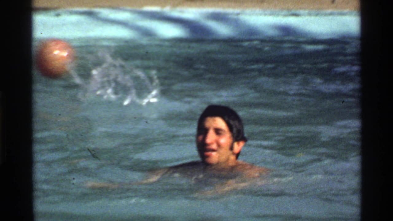 Adult man in a swimming pool near a small ball, with splashing water behind him.
