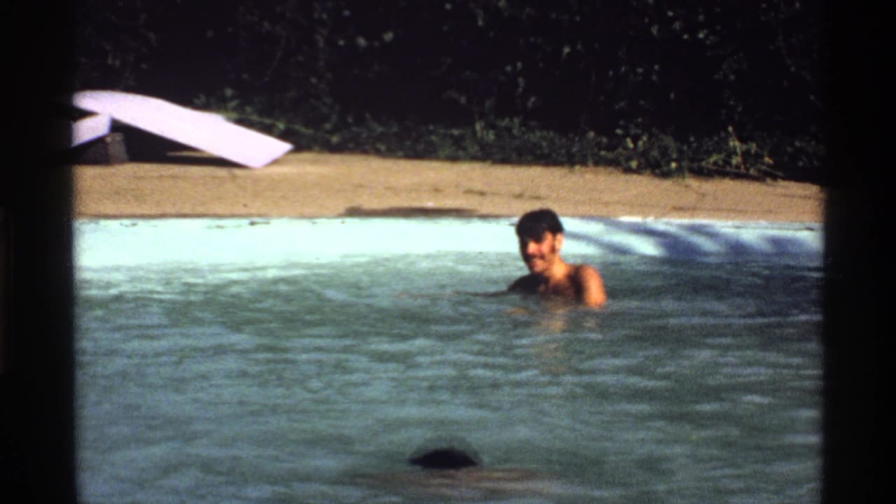 1978 California home-movie frame with a young man in a backyard pool, a white diving board, hedge, and pool edge behind him.