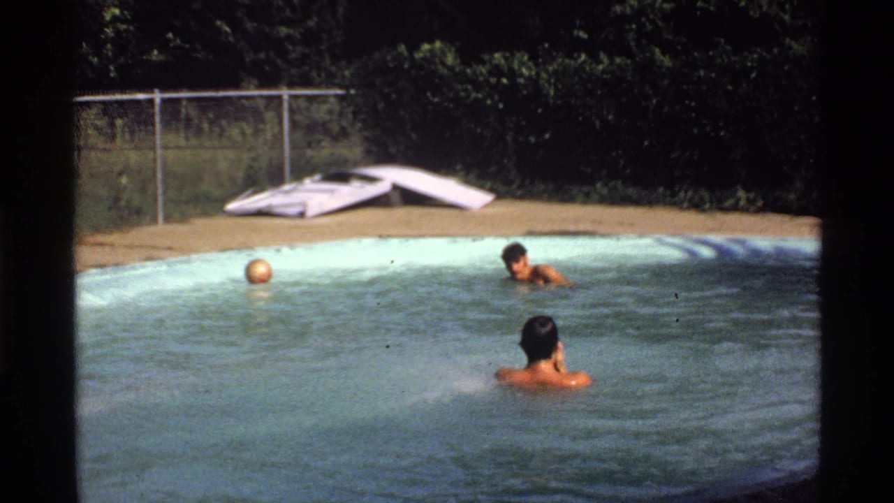 1978 California home-movie frame with two young men in the pool, a floating ball, chain-link fence, hedge, and white diving board.
