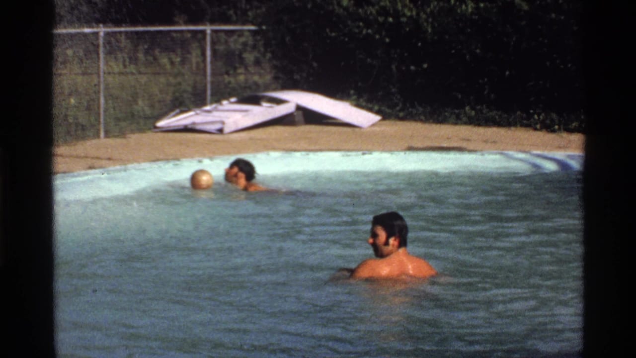1978 California home-movie frame with two adult men treading water in a backyard pool, a yellow ball nearby, and a chain-link fence in back.