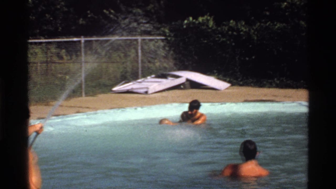 1978 California home-movie frame with three adult men in a backyard pool while a stream of water sprays across the scene from the left.