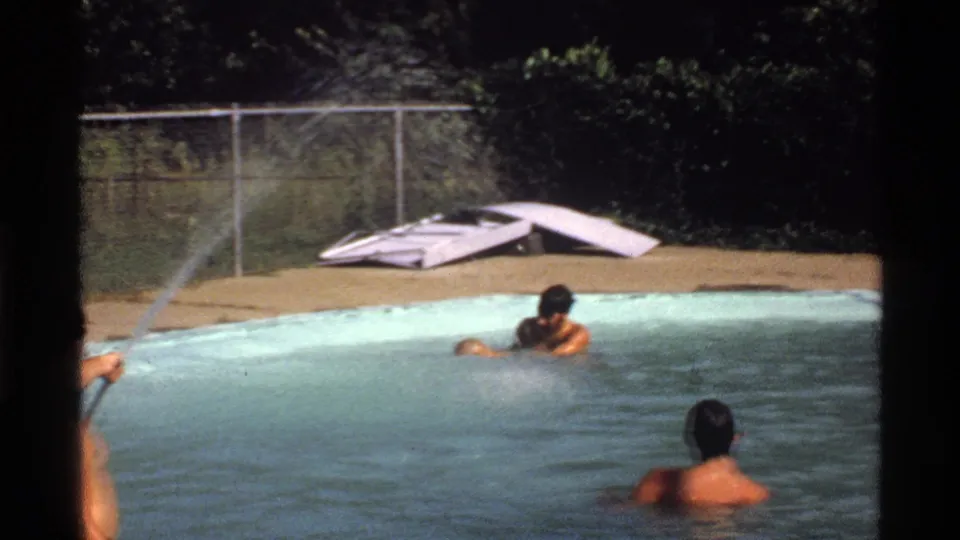 1978 California home-movie frame with three adult men in a backyard pool while a stream of water sprays across the scene from the left.