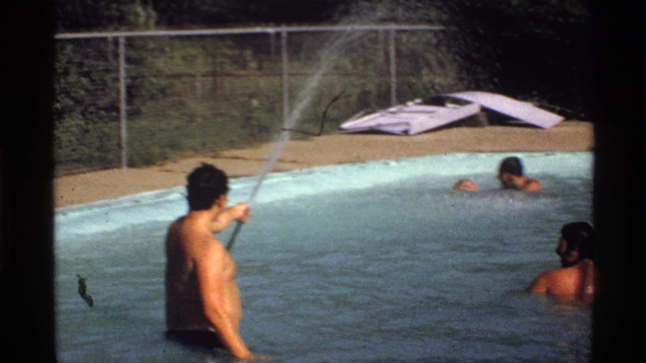 Man in a pool sprays a hose while two other men swim nearby.