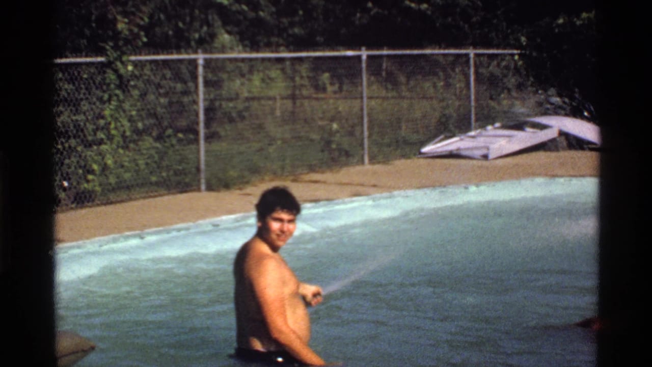 Man in swim trunks looks toward the camera while holding a hose in the pool.