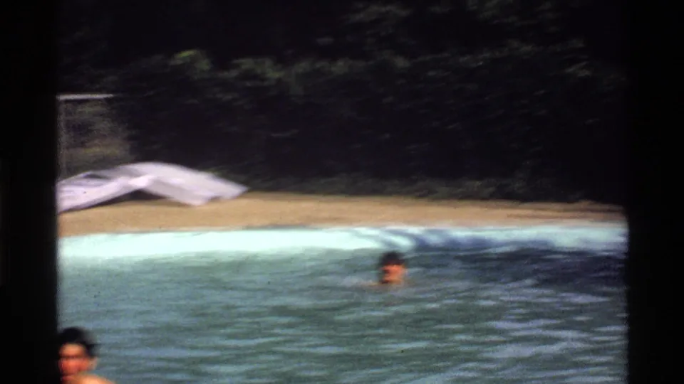 1978 California 8mm frame of swimmers partly visible in a backyard pool, with a white inflatable raft on the deck beyond them.