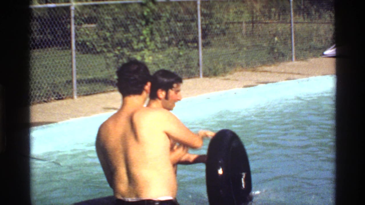 Two shirtless young men in a pool near a black inner tube and chain-link fence.