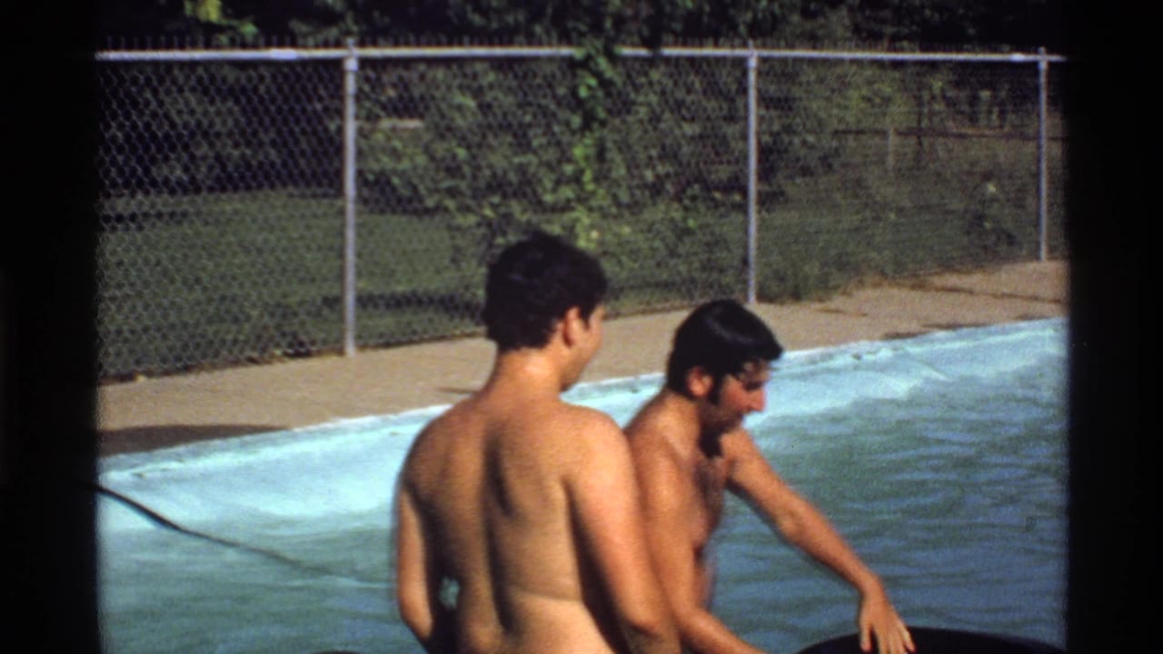 Two young men stand in pool water beside the pool edge and chain-link fence.