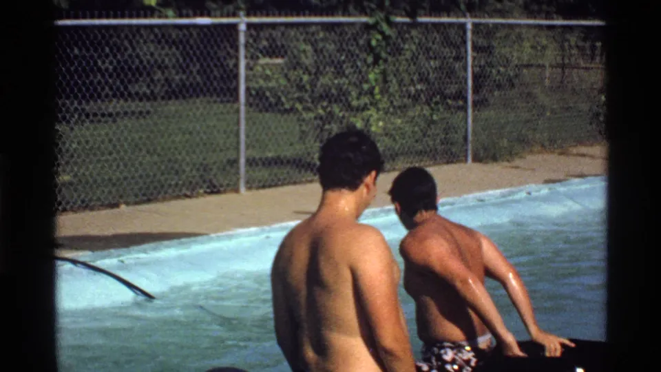 Two young men remain in the backyard pool, with one leaning on a black inner tube near the sunlit pool edge.