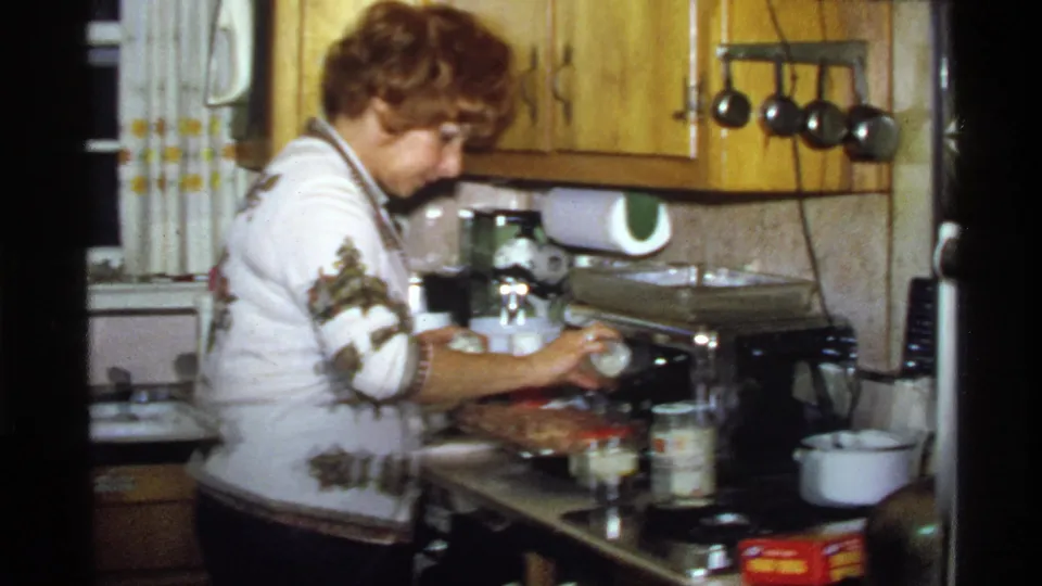 In a 1978 California home kitchen, a woman in a patterned sweater prepares food beside wood cabinets, jars, and a baking dish.