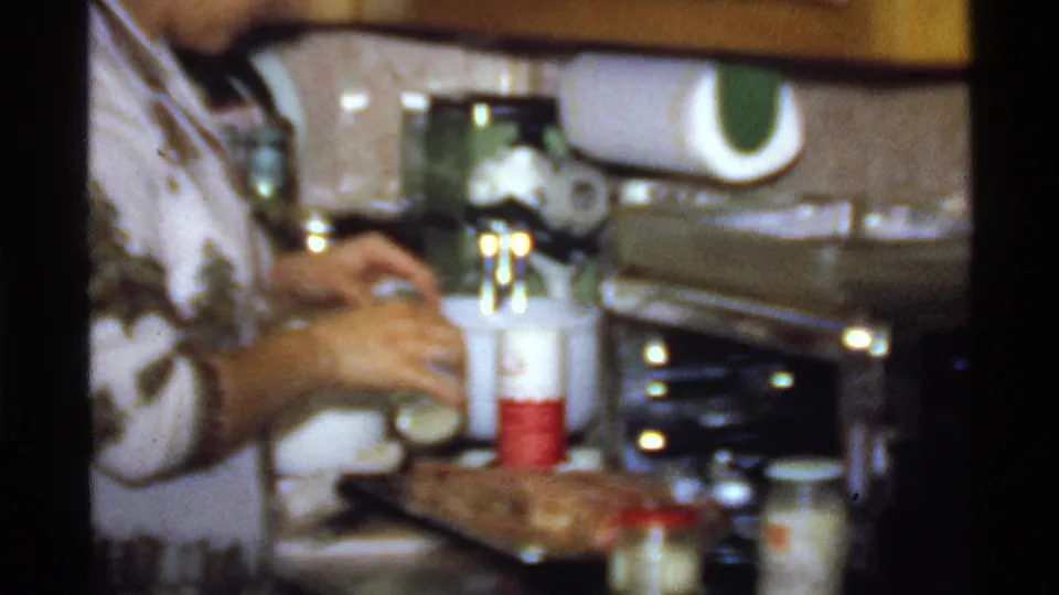 The woman continues seasoning a dish on the counter, with jars, appliances, cabinets, and a paper towel roll visible in the kitchen.
