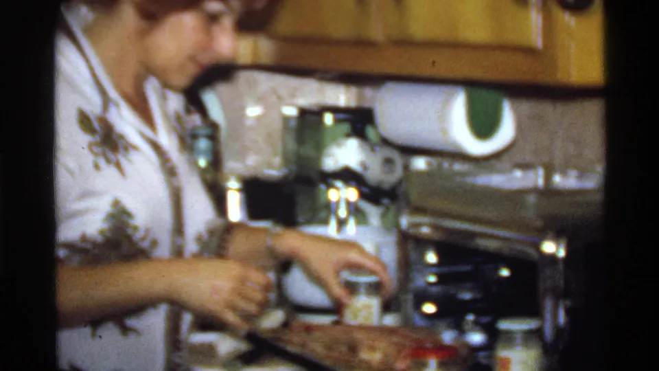 In the home kitchen, the woman reaches for or opens a jar beside a seasoned dish, with countertop appliances and cabinets behind her.