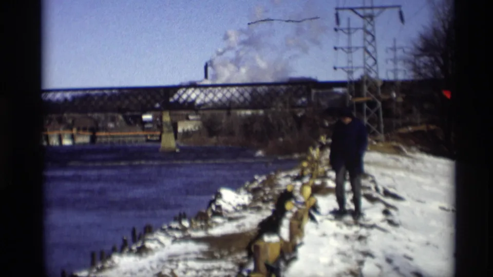 A distant adult walks along a snowy riverbank in a Wisconsin home-movie scene, with a bridge, utility tower, and industrial steam beyond the water.