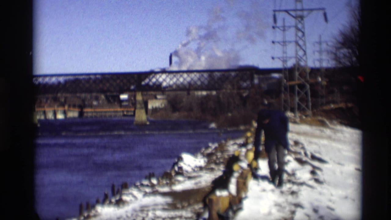 Person in a dark coat walking along a snowy riverbank near a steel bridge.