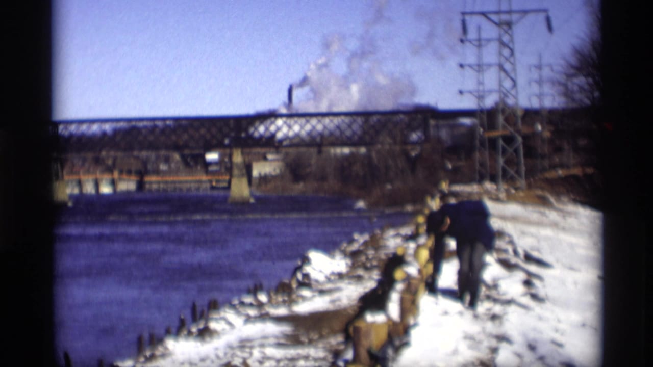 1966 Madison frame showing a person moving along a snow-covered river edge, with a truss bridge, utility tower, and distant smoke visible.