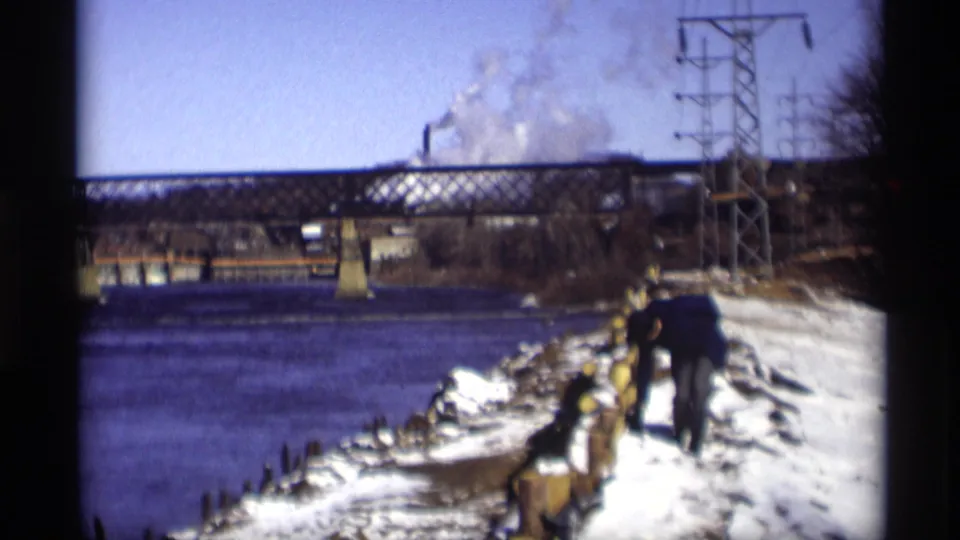 1966 Madison frame showing a person moving along a snow-covered river edge, with a truss bridge, utility tower, and distant smoke visible.
