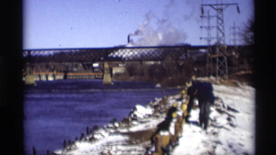 Madison, Wisconsin, 1966 frame of a dark-coated figure on a snowy riverbank below a steel bridge and winter industrial skyline.