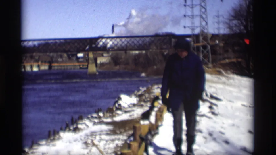 1966 Madison home-movie frame with the walker closer to camera on a snowy bank, river to the left, bridge and power tower in the background.
