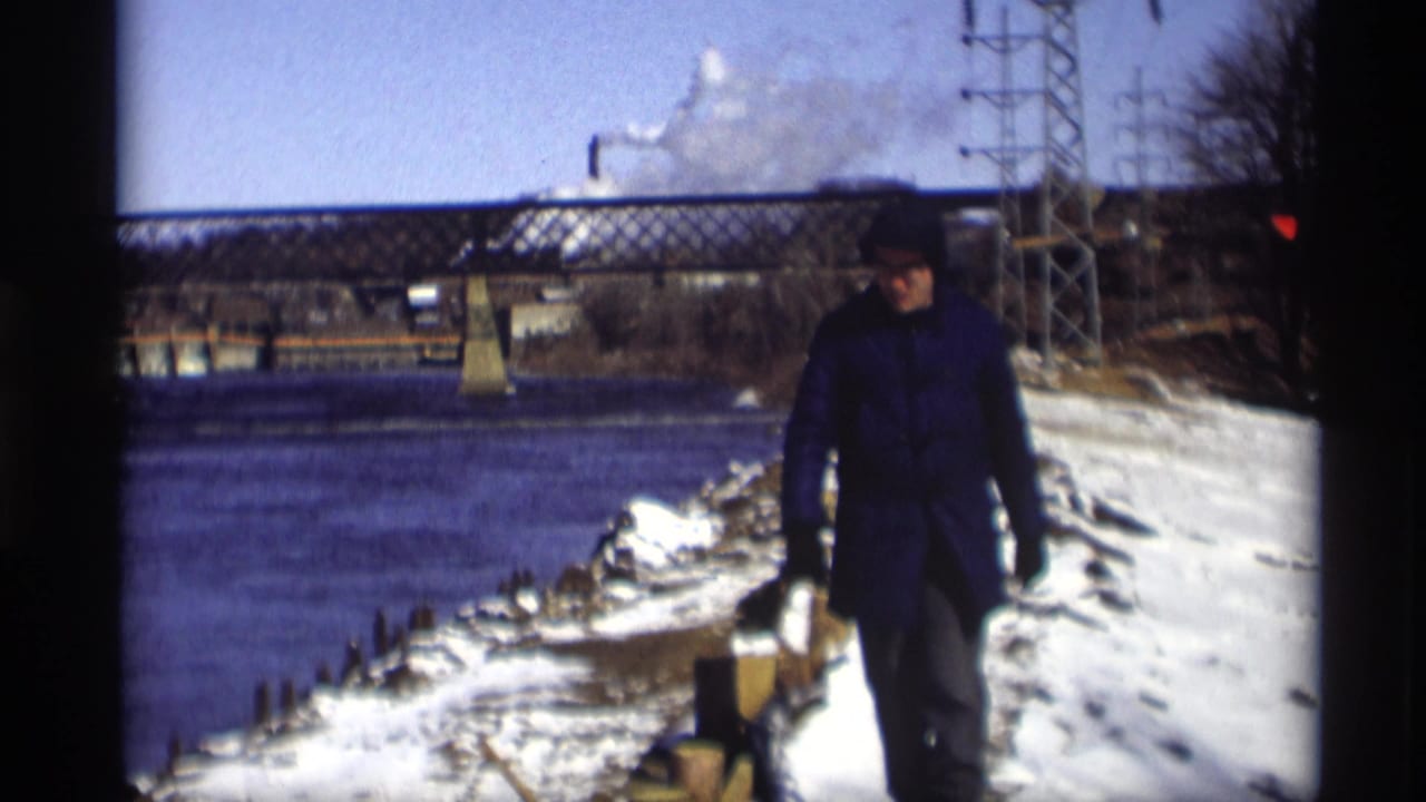 In Madison, Wisconsin, a man walks along a snow-covered riverbank with river water, a truss bridge, utility towers, and smoke visible behind him.