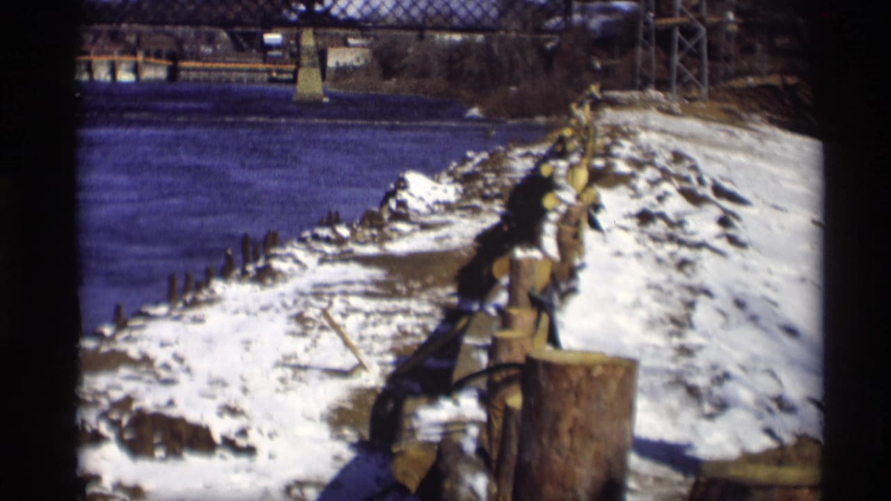 A Madison riverbank in winter shows snow, wooden shoreline pilings, dark river water, a distant truss bridge, and industrial smoke beyond the bank.