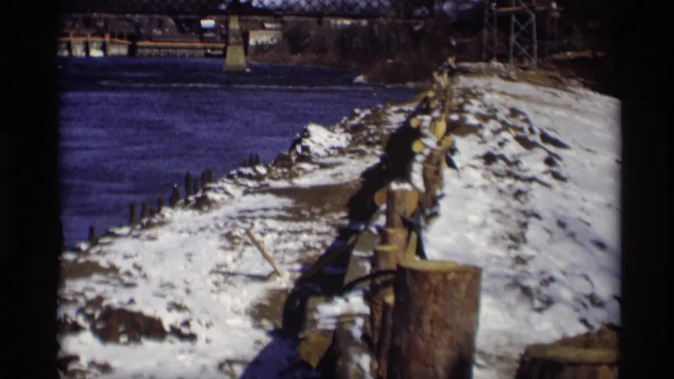 The winter shoreline continues along the river, with snow, rough wooden pilings, dark water, and a truss bridge visible in the Madison background.