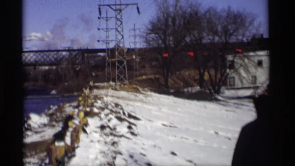 The Madison winter riverbank is seen from a wider angle, showing snow, utility towers, bare trees, a truss bridge, and a small light-colored structure.