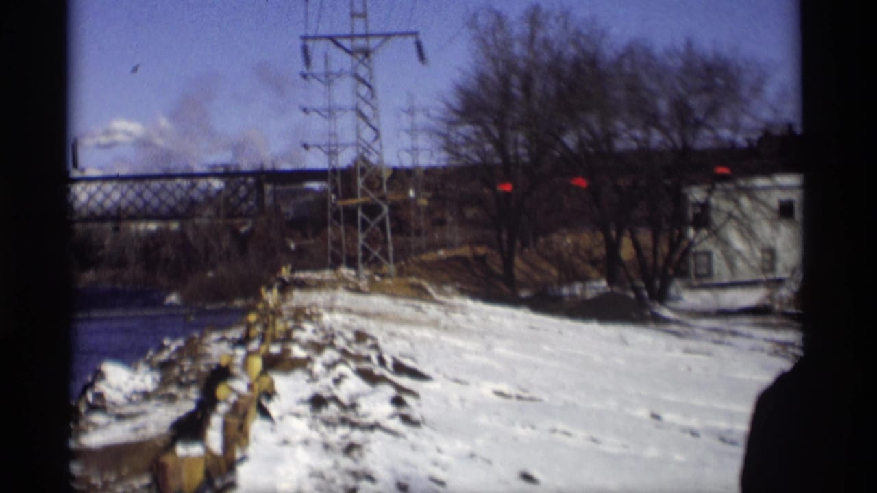 Snowy riverbank with a power-line tower, utility lines, trees, and distant structures.