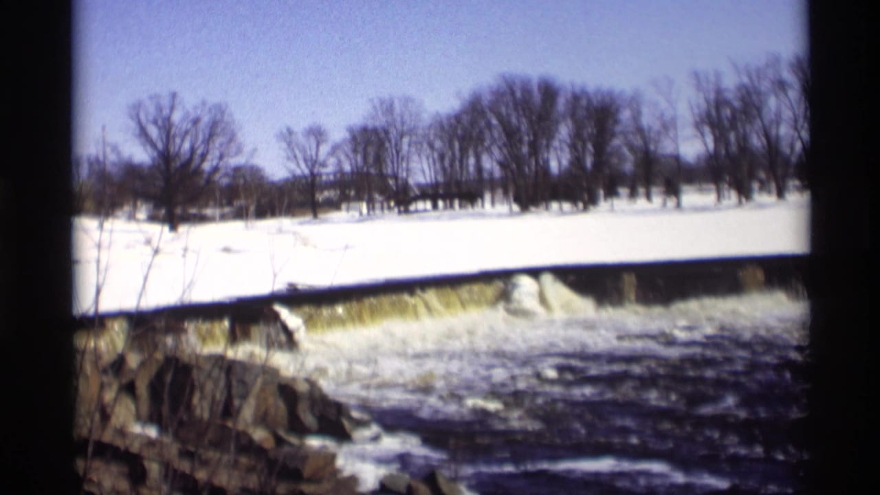 1966 Madison 8mm frame of a low river dam in winter, with foaming water, snow-covered banks, rocks, and bare trees.