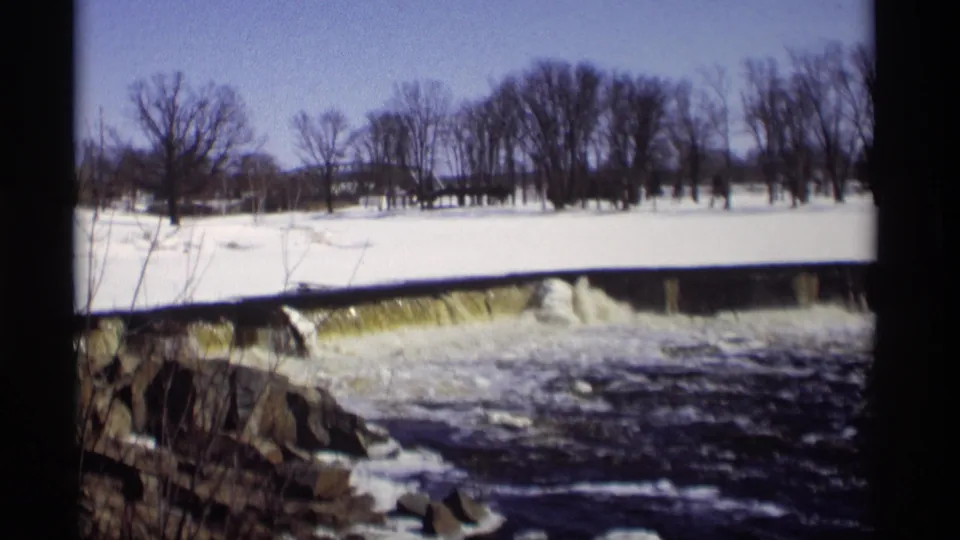 1966 Madison winter frame showing a snowy open bank, low dam, rocky foreground, rushing water, and leafless tree line.