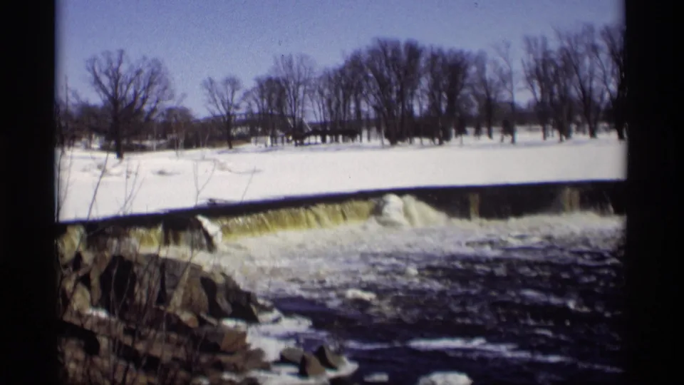 1966 Madison 8mm frame of a low dam and dark turbulent river water bordered by snow, rocks, and bare winter trees.