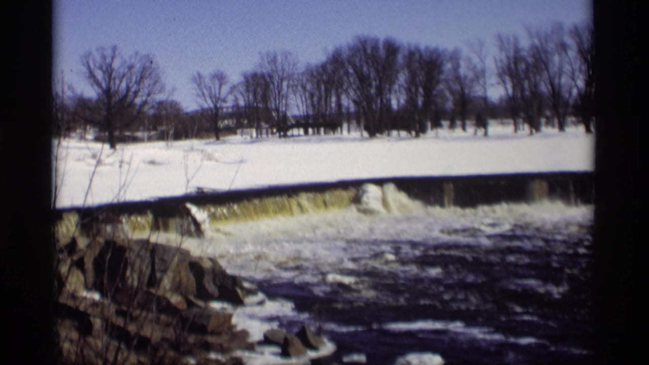 1966 Madison, Wisconsin 8mm frame showing fast water spilling over a low dam in a snowy winter landscape with bare trees.
