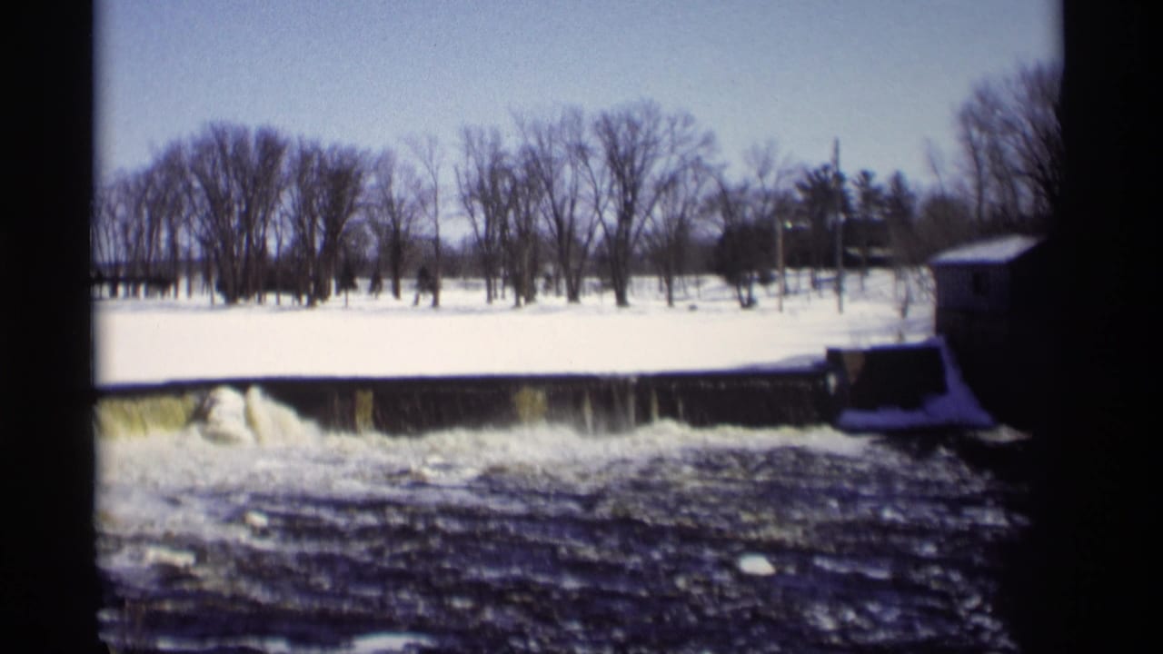 The pan reveals more of the snowy dam scene in Madison, Wisconsin, with turbulent water, winter trees, and a small riverside structure.